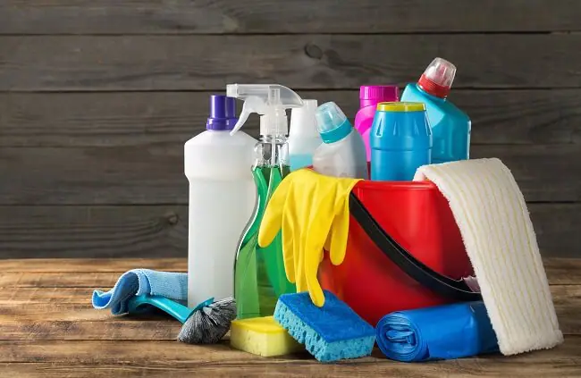 A red bucket sits on top of a wood table top. The bucket is full of cleaning supplies and supplies sit beside it too.